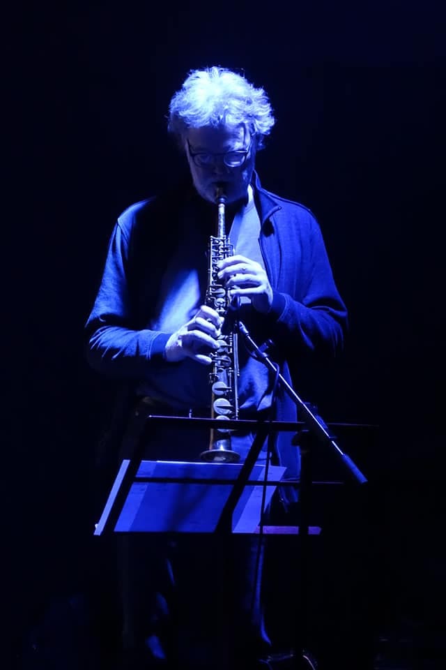 A person is playing a wind instrument under blue stage lighting, standing in front of a music stand.