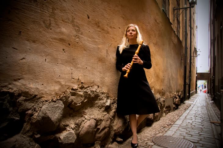 A woman in a black dress stands in a narrow alley holding a wooden recorder.