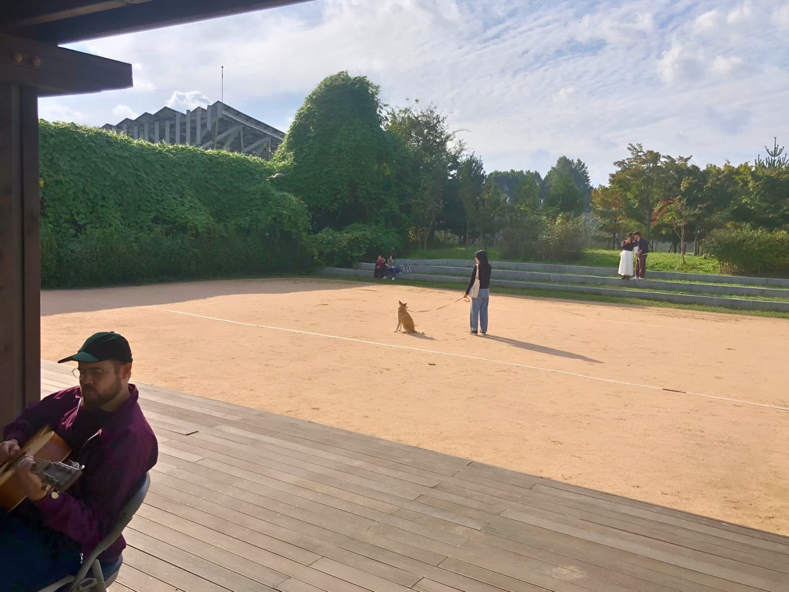 A person plays guitar under a shelter by a sandy area, where another person is walking a dog, with greenery and people in the background.