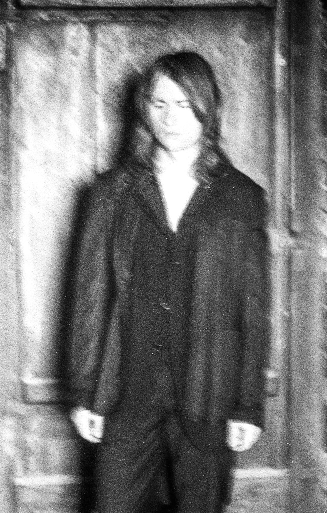 A blurry black-and-white photo of a person with long hair wearing a suit, standing against a textured background.
