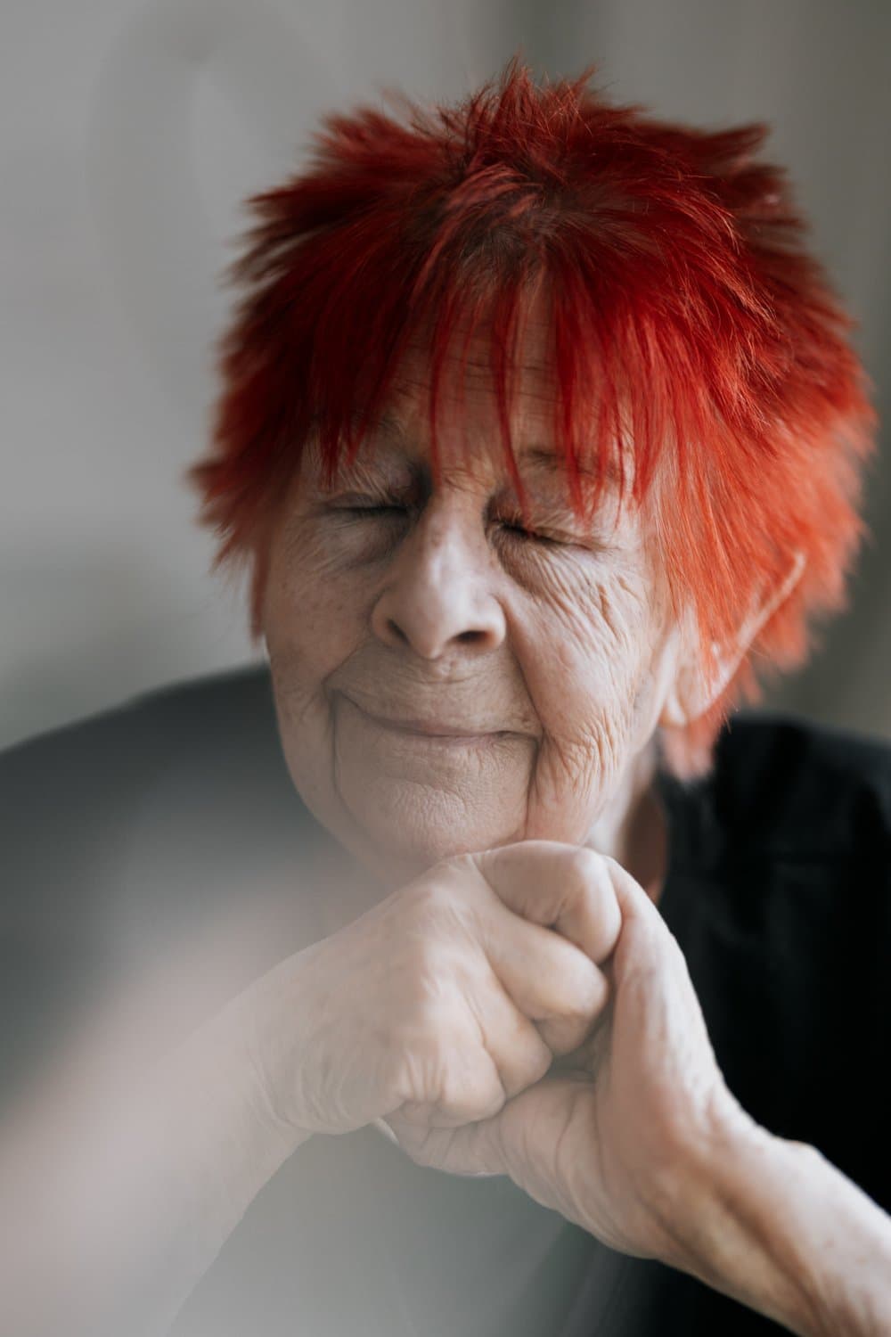 An elderly person with bright red hair is smiling with their eyes closed and hands clasped under their chin.