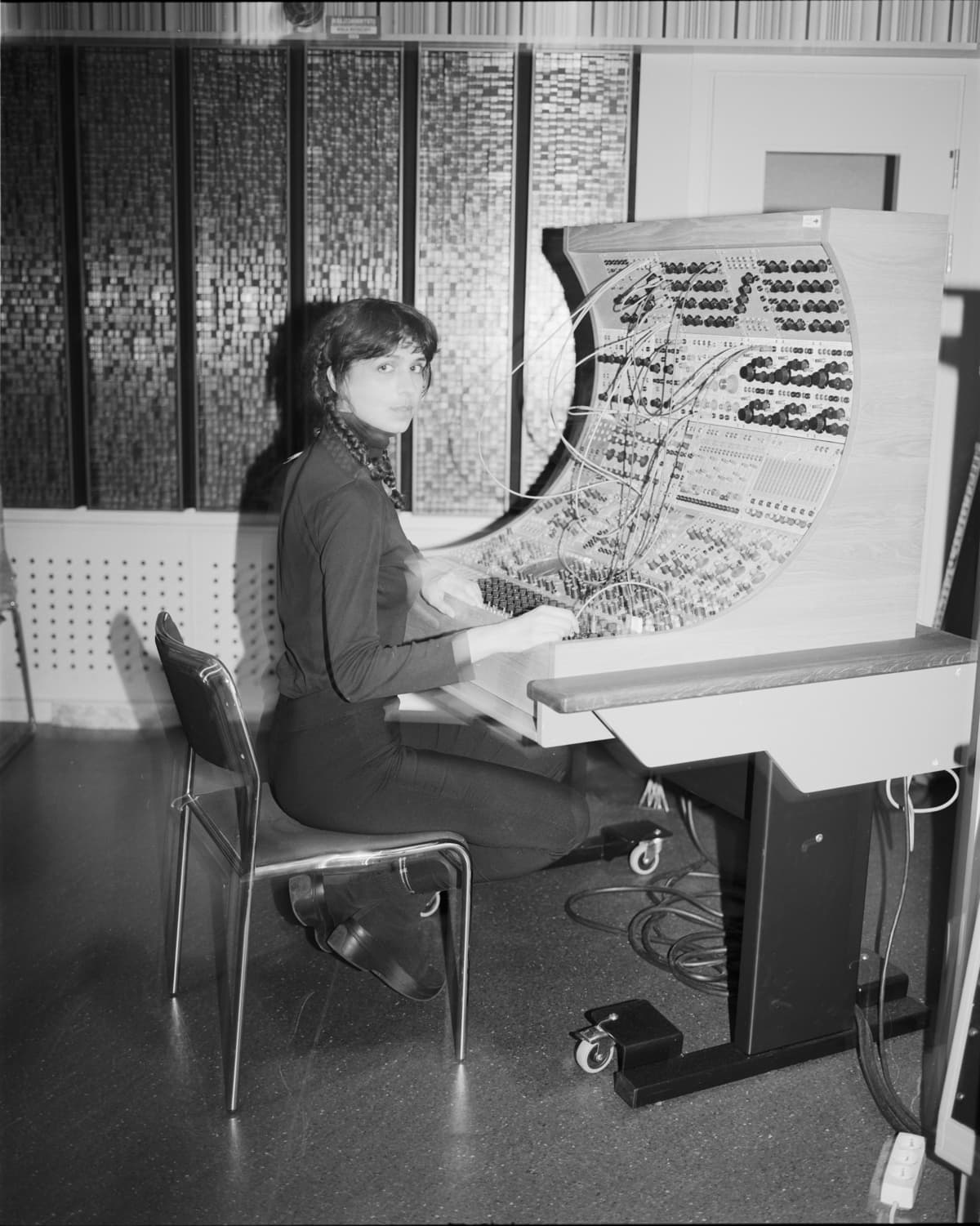 A person is seated at an early analog synthesizer with numerous knobs, dials, and patch cables in a studio setting.