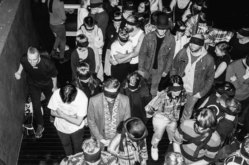 A group of people, many of whom are wearing blindfolds, are gathered closely together in what appears to be an indoor space.
