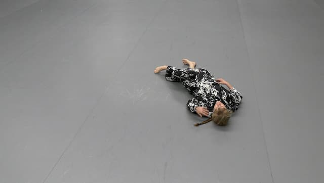 A person in a black and white patterned outfit is lying on their side on a gray floor, possibly a dance or martial arts studio.