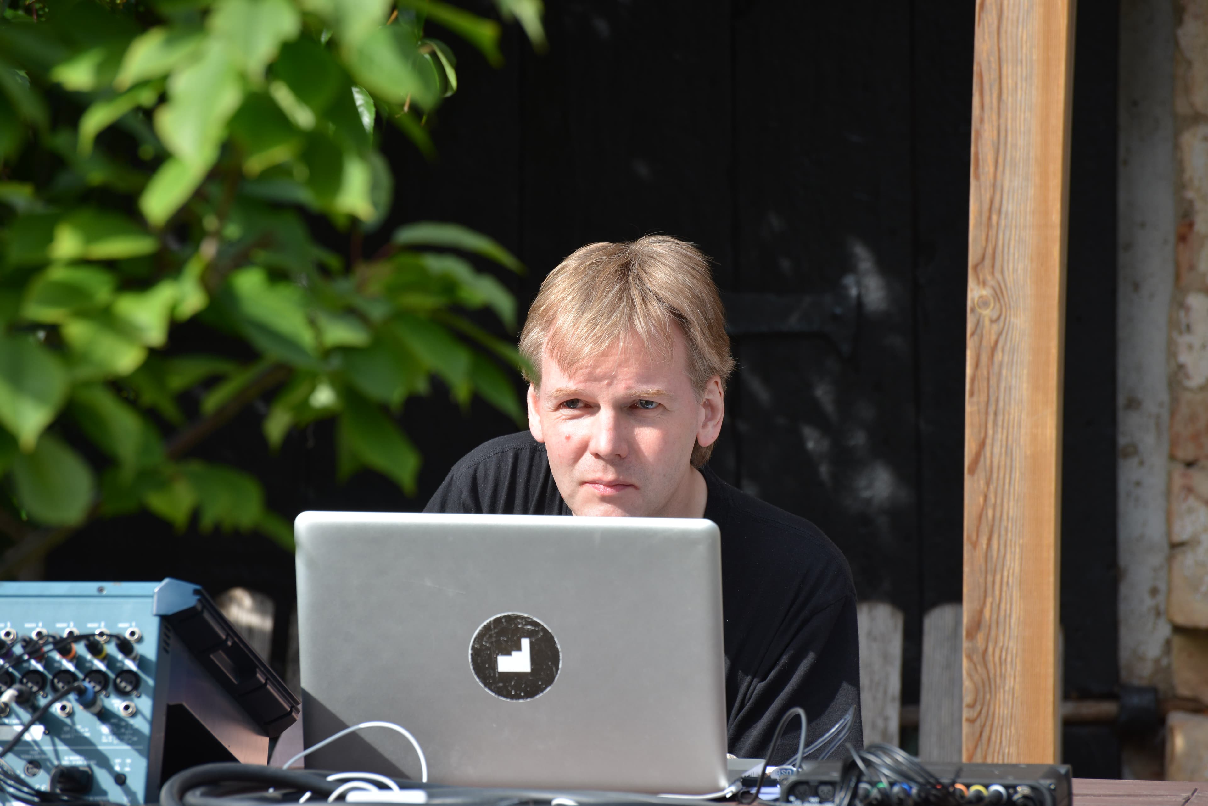 A person with blond hair is sitting outdoors, looking at a laptop with various electronic equipment nearby.