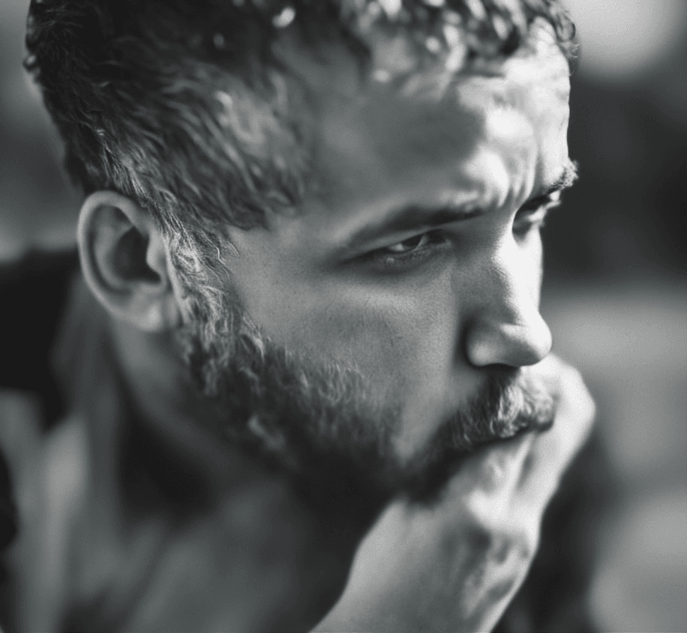 A black-and-white close-up of a man with a beard and short hair, looking pensively to the side while touching his chin.