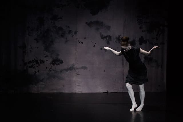 A person dressed in black with white makeup performs a dramatic dance on a dark stage with a textured backdrop.