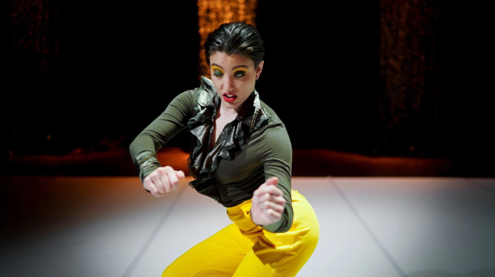 A performer with dramatic makeup and bright yellow pants strikes a dynamic pose on stage.