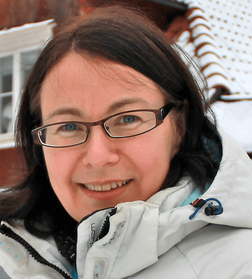 A woman wearing glasses and a winter jacket smiles outdoors with a snowy background.