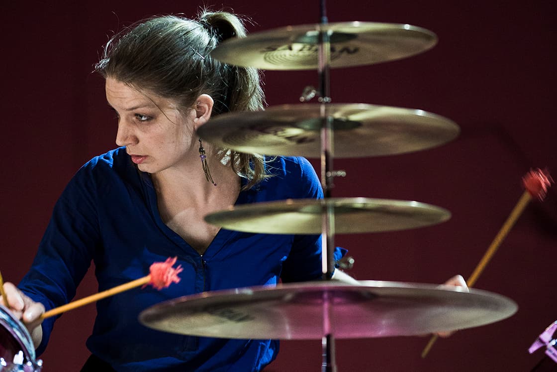 A person in a blue shirt is playing drums with mallets, with a focus on the cymbals in the foreground.