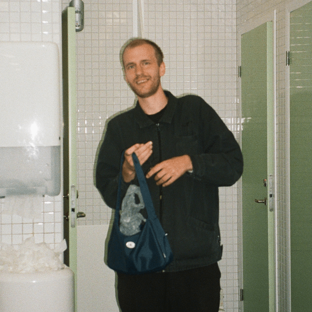 A person stands in a restroom with white tiled walls, holding a blue bag, and smiling, with a messy paper towel dispenser in the background.