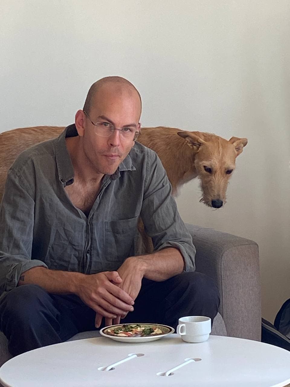 A man with glasses and a dog sit on a couch, with the man eating from a plate on a table that also has a cup and two spoons.