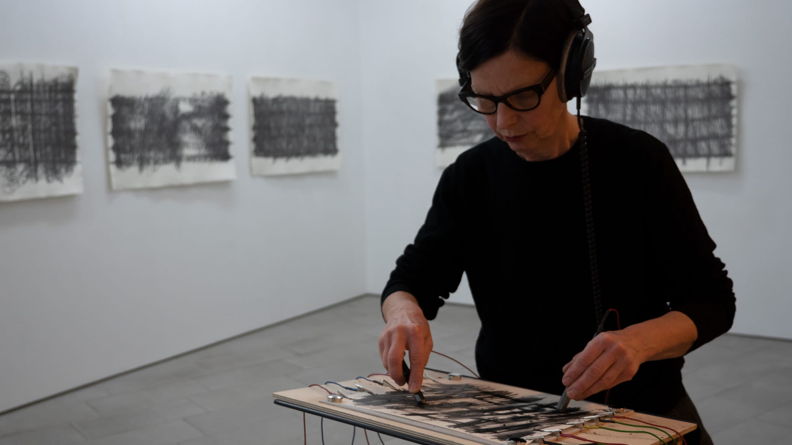 A person wearing headphones and glasses is drawing with charcoal on paper connected to wires, with abstract artworks displayed on the wall behind them.