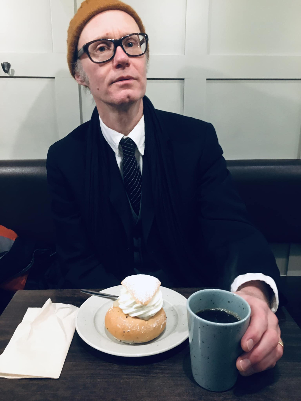 Kostymklädd man vid ett kafébord med kaffekopp och semla.